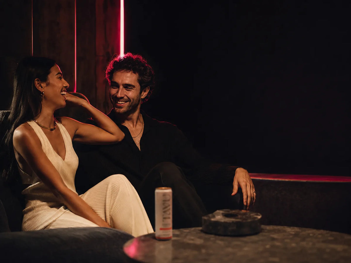 Two people mid-laugh on a sofa, shoulders relaxed, a TAVU can on the table